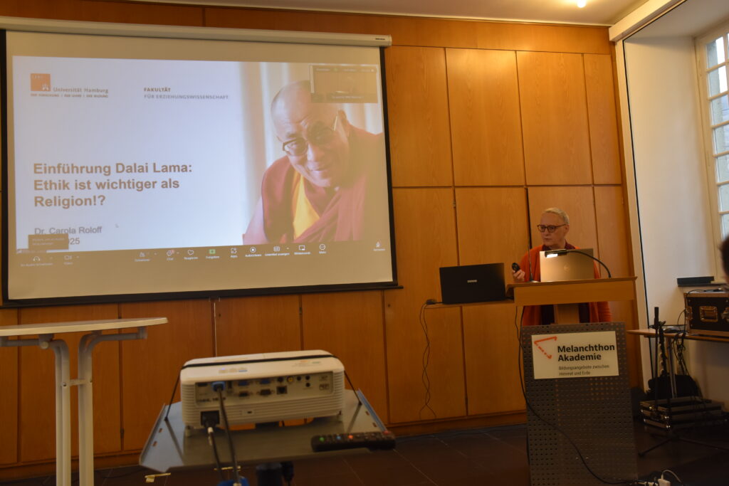 Das Bild zeigt Carola Roloff am Rednerpult und eine PowerPoint-Folie mit dem Dalai Lama, Text: Einführung Dalai Lama: Ethik wichtiger als Religion?"