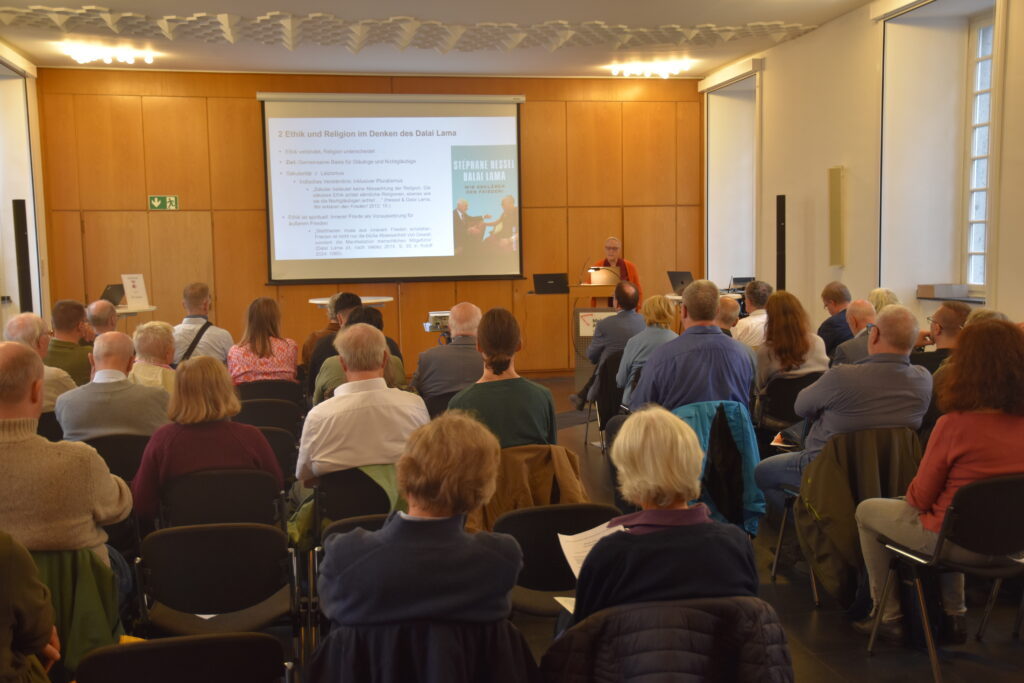 Das Bild zeigt ca 30 Zuhörerinnen und Zuhörer von hinten. Sie schauen auf eine Leinwand mit einem Powerpointvortrag. Carola Roloff steht am Rednerpult