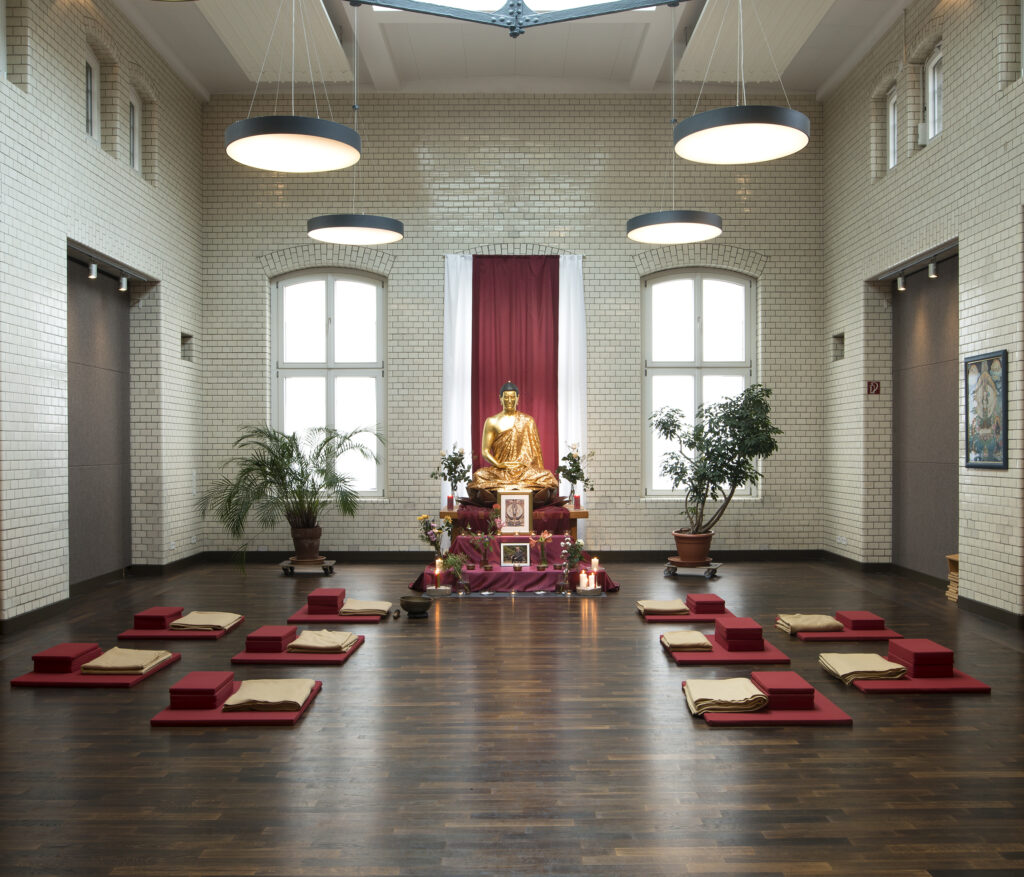 Schreinraum mit einer goldenen Buddha-Statue, gerahmt von 2 großen Grünpflanzen und 10 Meditationskissen in Rot davor. 